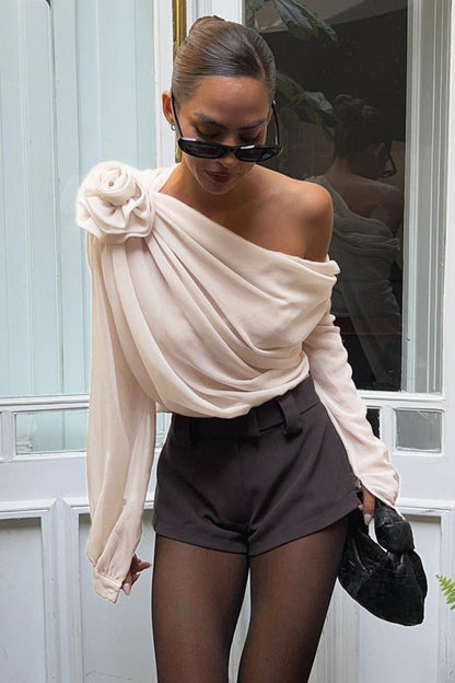 Beige - A woman wearing an elegant, cream-coloured, one-shoulder draped blouse featuring a large 3D floral appliqué on the right shoulder and a single long sleeve.