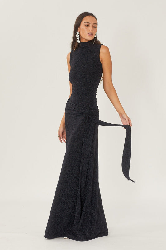 Black - A woman wearing a formal, floor-length black sleeveless maxi dress featuring a high neckline, a ruched tie-waist detail, and a subtle shimmering finish.