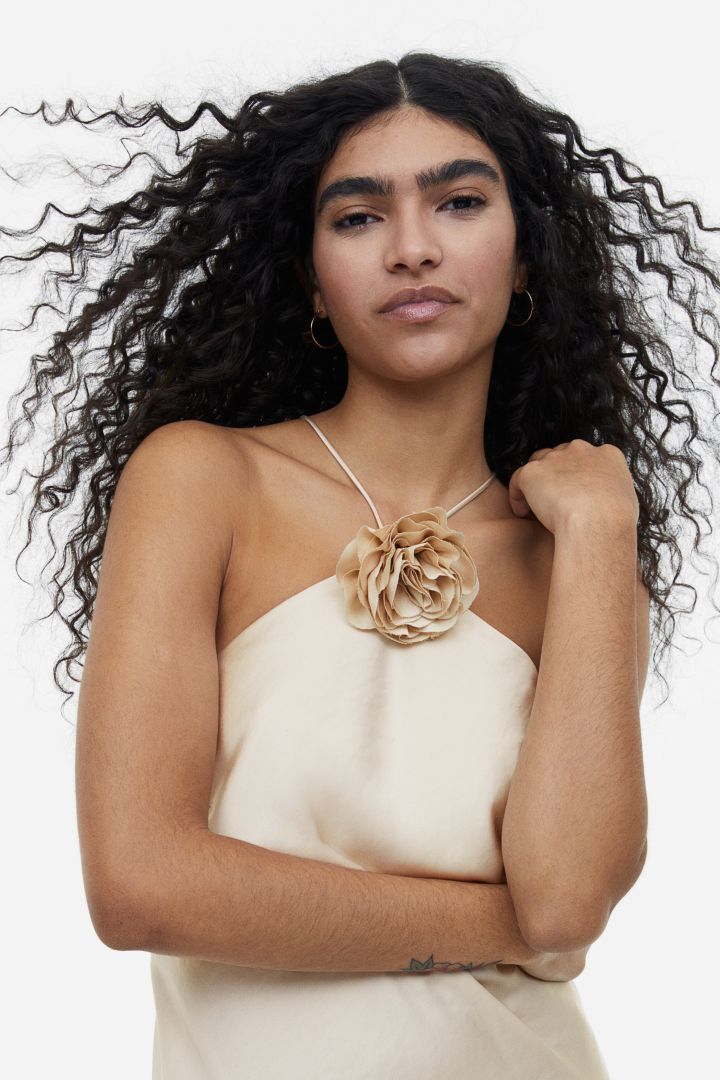 Beige - A woman wearing a pale champagne, satin-finish maxi slip dress featuring a halter neckline with a large 3D floral appliqué.