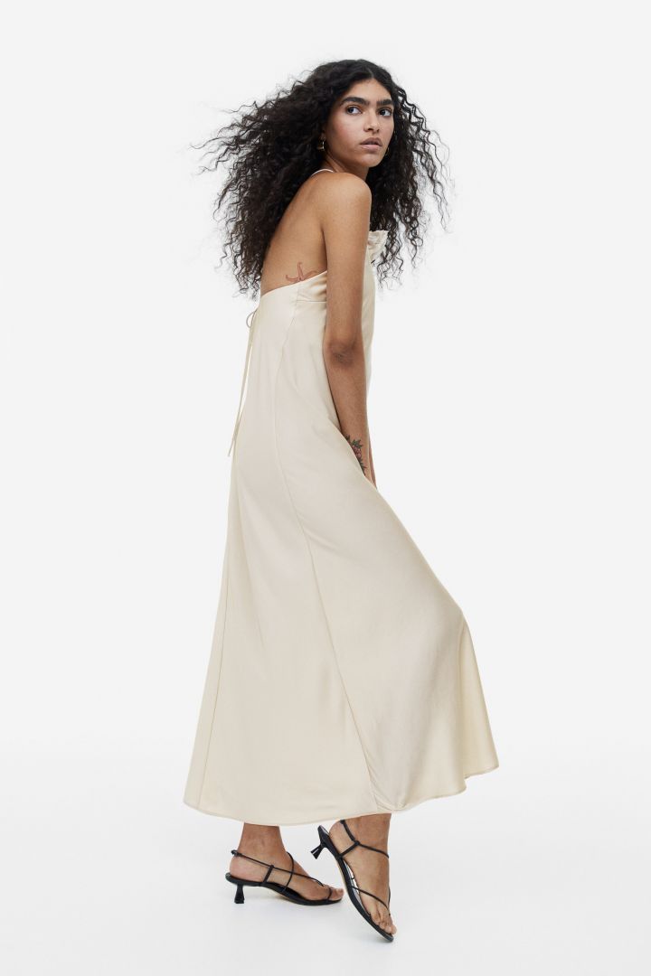 Beige - A woman wearing a pale champagne, satin-finish maxi slip dress featuring a halter neckline with a large 3D floral appliqué.