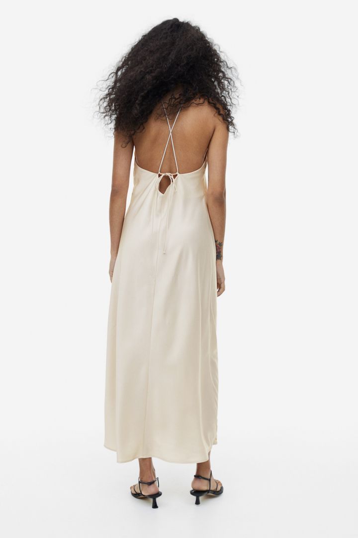 Beige - A woman wearing a pale champagne, satin-finish maxi slip dress featuring a halter neckline with a large 3D floral appliqué.