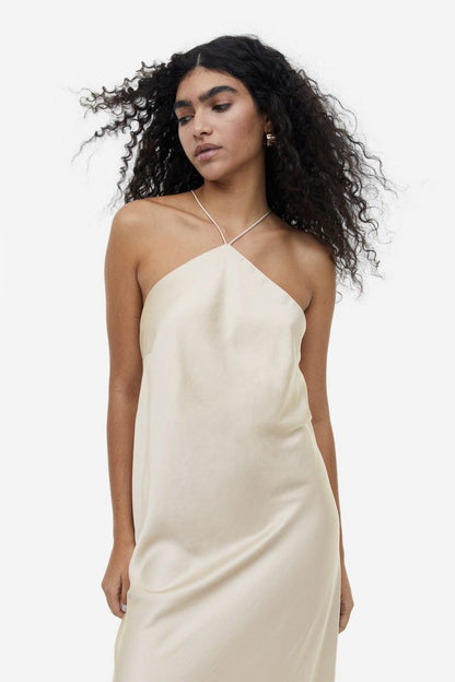 Beige - A woman wearing a pale champagne, satin-finish maxi slip dress featuring a halter neckline with a large 3D floral appliqué.