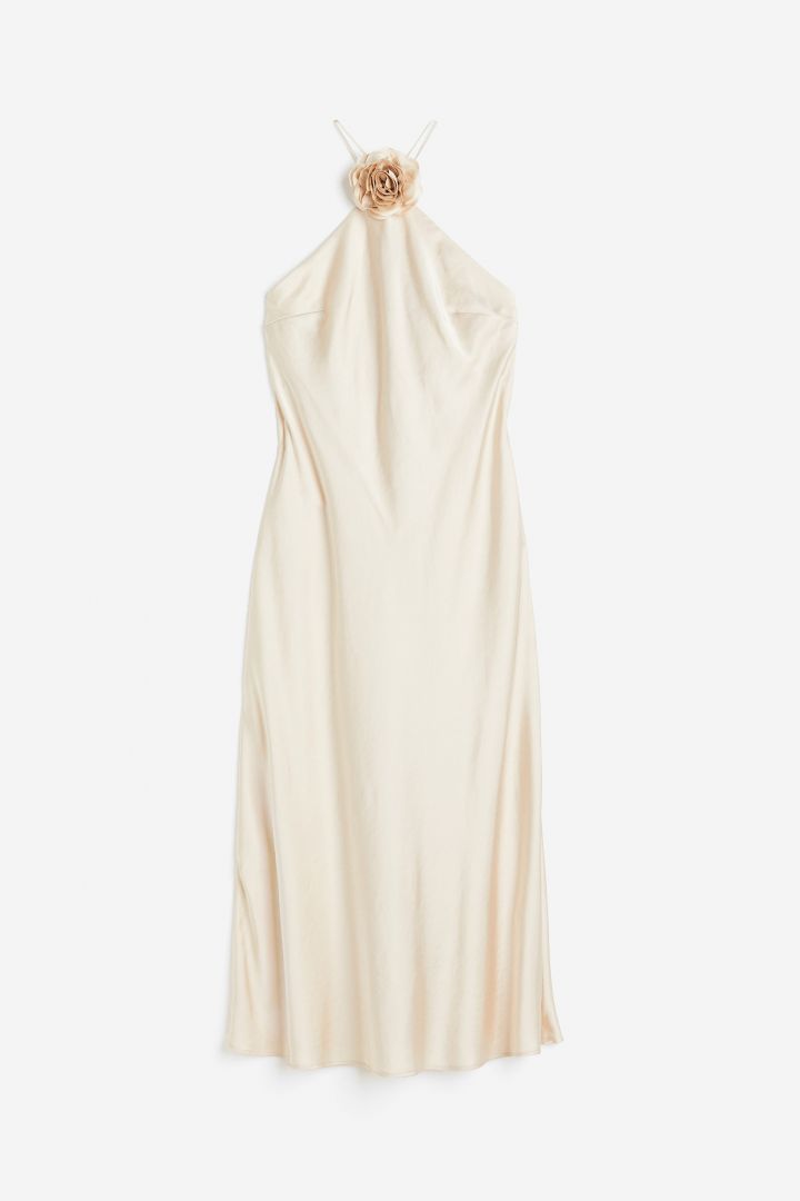 Beige - A woman wearing a pale champagne, satin-finish maxi slip dress featuring a halter neckline with a large 3D floral appliqué.