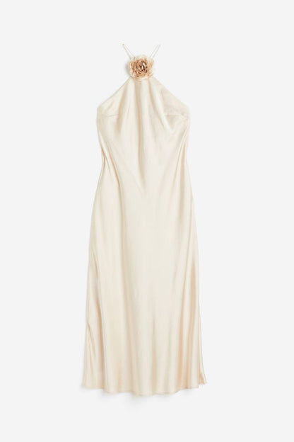 Beige - A woman wearing a pale champagne, satin-finish maxi slip dress featuring a halter neckline with a large 3D floral appliqué.