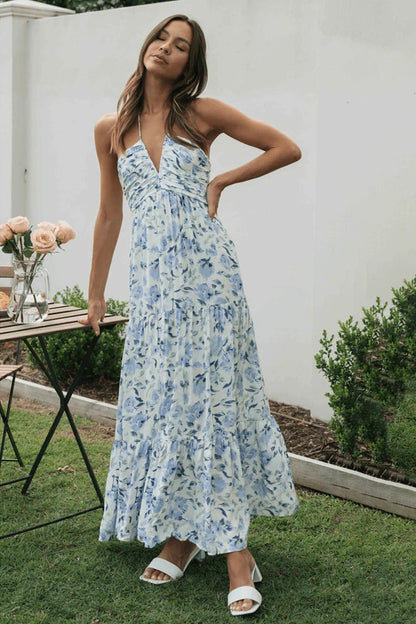 Blue - A woman wearing a white maxi dress with an orange and blue floral print, featuring a halter neckline, ruched bodice, and tiered skirt, standing outdoors.