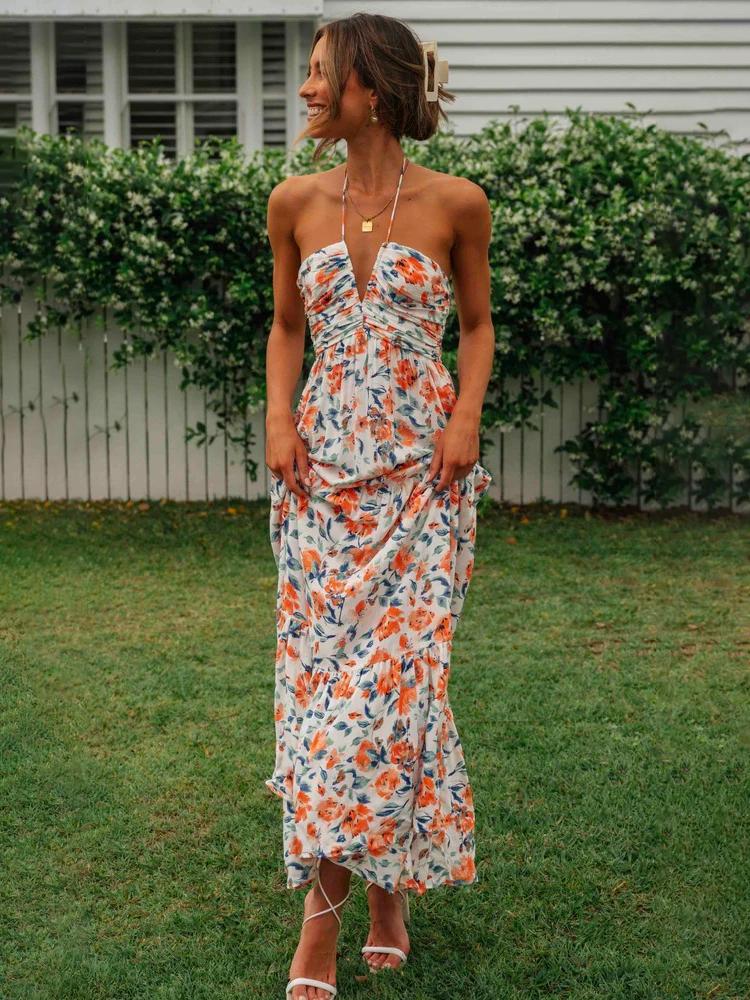 Orange - A woman wearing a white maxi dress with an orange and blue floral print, featuring a halter neckline, ruched bodice, and tiered skirt, standing outdoors.