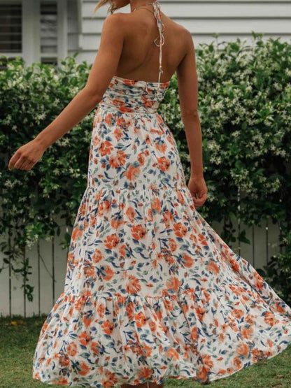 Orange - A woman wearing a white maxi dress with an orange and blue floral print, featuring a halter neckline, ruched bodice, and tiered skirt, standing outdoors.