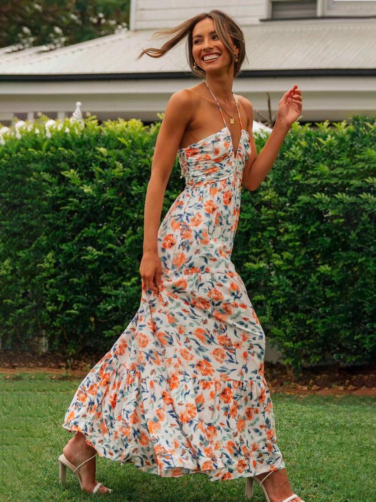 Orange - A woman wearing a white maxi dress with an orange and blue floral print, featuring a halter neckline, ruched bodice, and tiered skirt, standing outdoors.