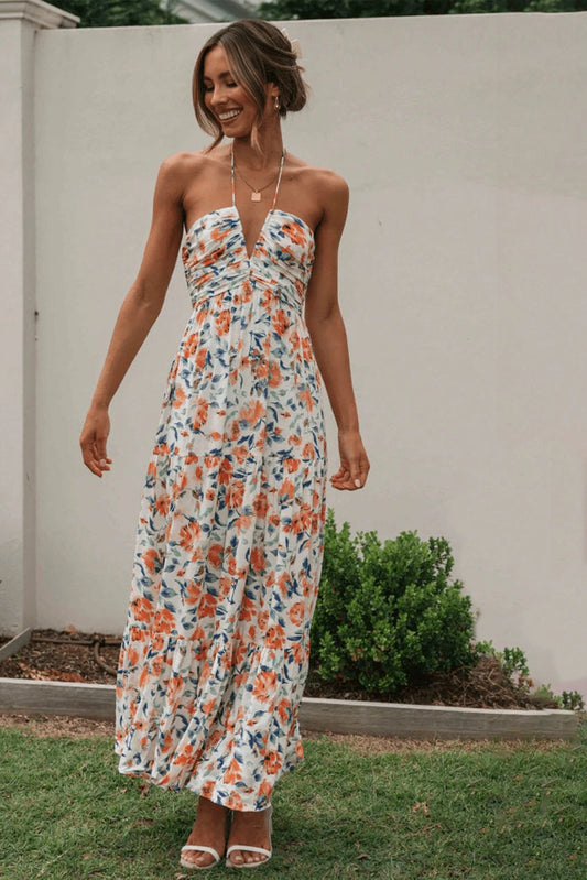 Orange - A woman wearing a white maxi dress with an orange and blue floral print, featuring a halter neckline, ruched bodice, and tiered skirt, standing outdoors.