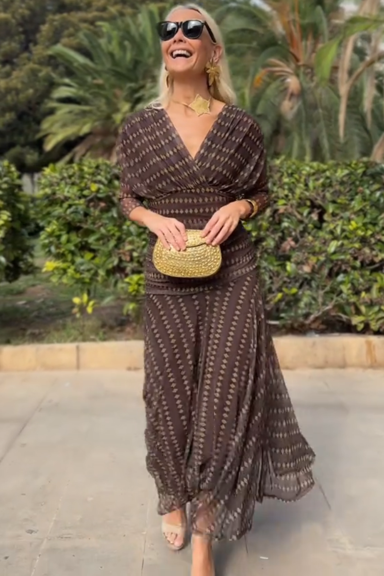 Brown - A woman smiling outdoors wearing an elegant, flowing brown maxi dress with a geometric diamond print, featuring a wrap-style V-neckline, sheer 3/4 sleeves, and a full skirt.