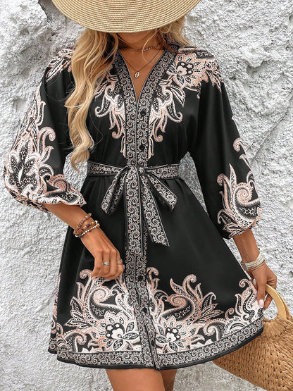 Black - Woman wearing a woven boho V-neck mini shirt dress with ornate paisley print and voluminous sleeves, holding a woven straw bag outdoors.