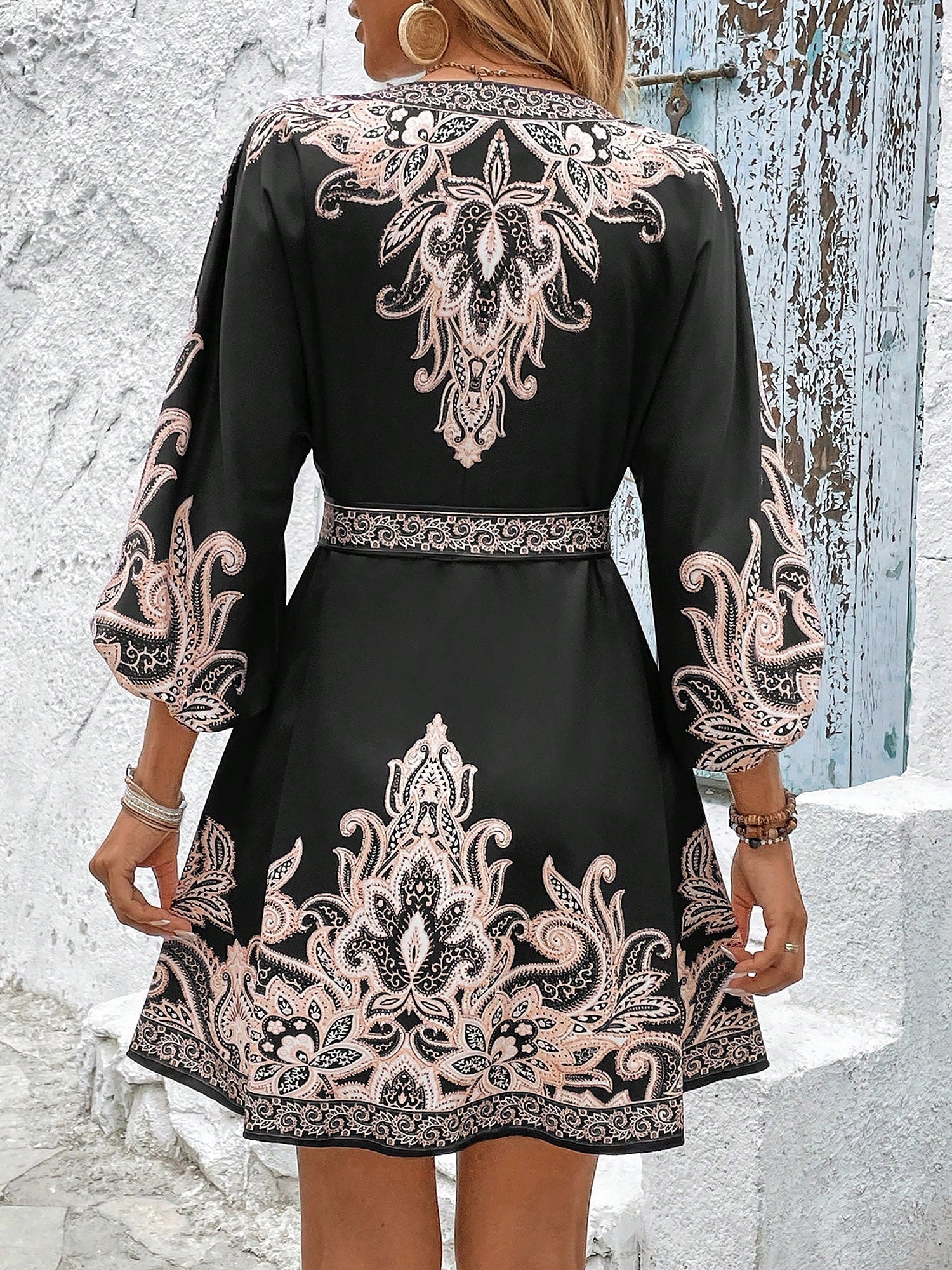Black - Woman wearing a woven boho V-neck mini shirt dress with ornate paisley print and voluminous sleeves, holding a woven straw bag outdoors.