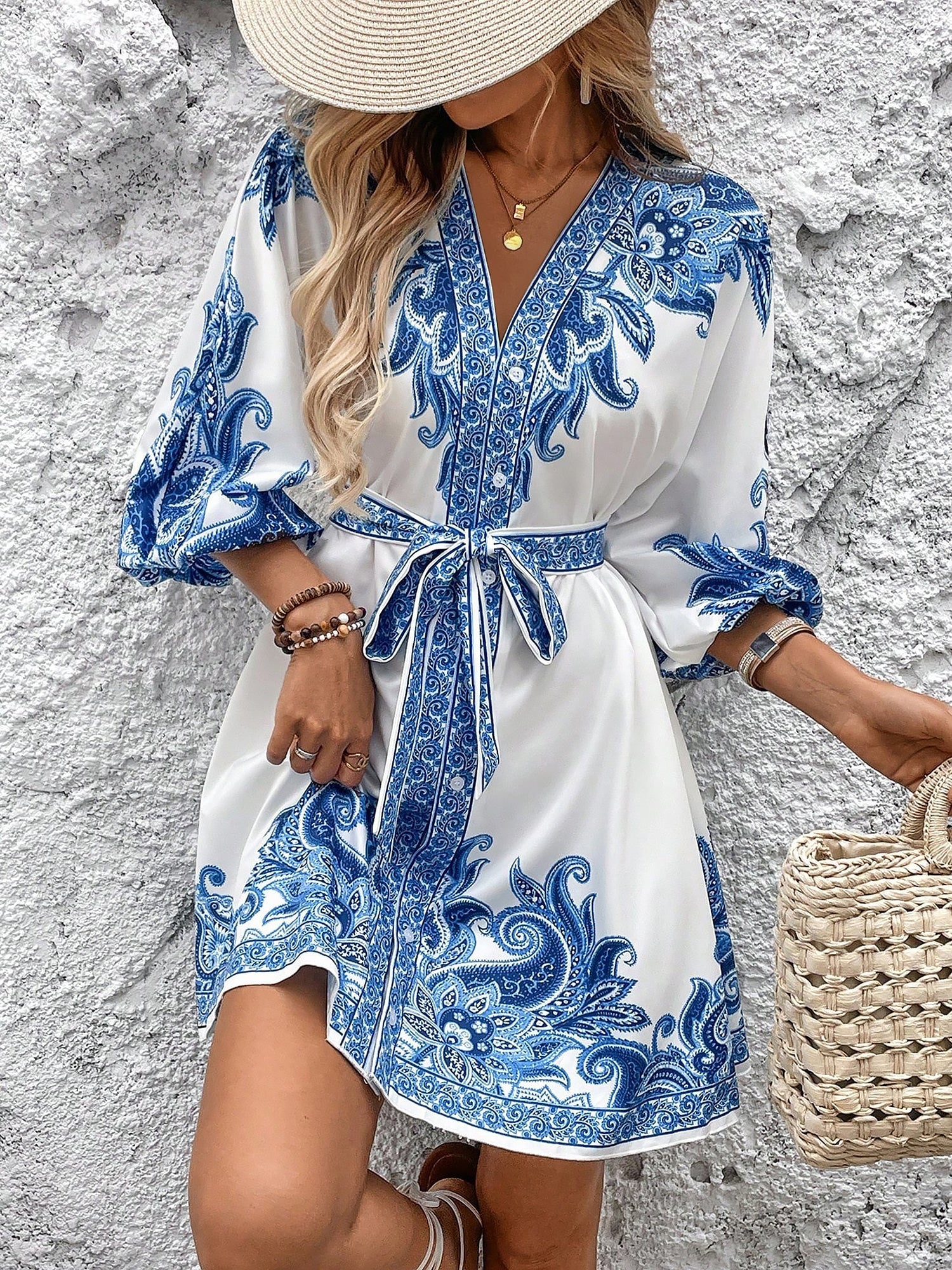 Blue - Woman wearing a woven boho V-neck mini shirt dress with ornate paisley print and voluminous sleeves, holding a woven straw bag outdoors.