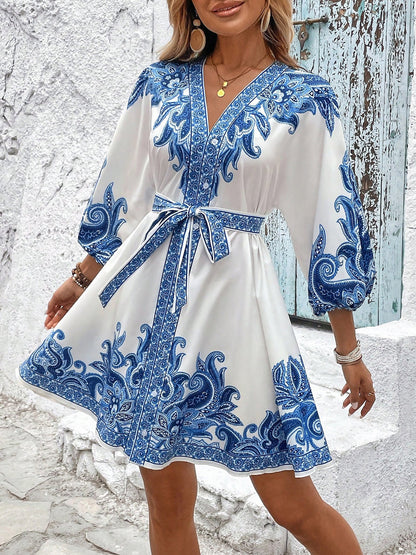 Blue - Woman wearing a woven boho V-neck mini shirt dress with ornate paisley print and voluminous sleeves, holding a woven straw bag outdoors.