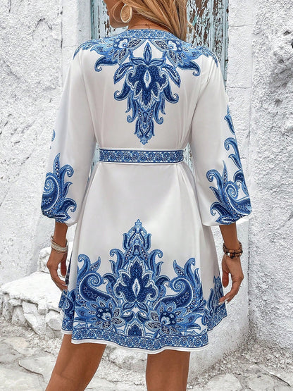 Blue - Woman wearing a woven boho V-neck mini shirt dress with ornate paisley print and voluminous sleeves, holding a woven straw bag outdoors.