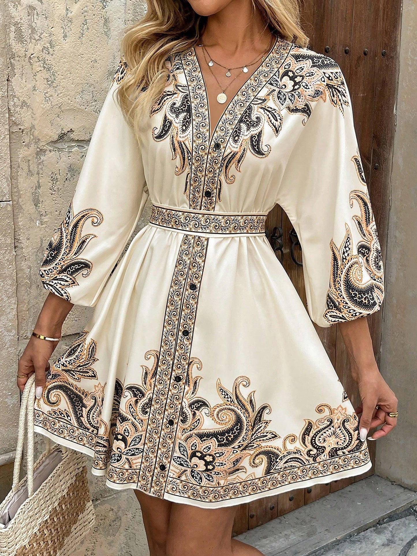 Apricot - Woman wearing a woven boho V-neck mini shirt dress with ornate paisley print and voluminous sleeves, holding a woven straw bag outdoors.