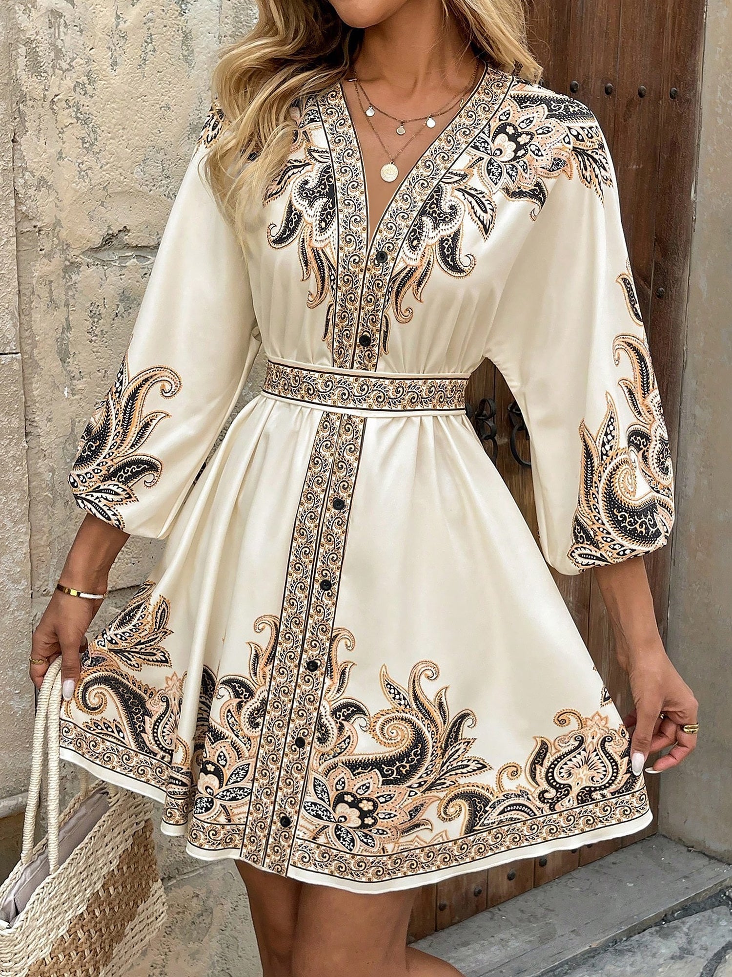 Apricot - Woman wearing a woven boho V-neck mini shirt dress with ornate paisley print and voluminous sleeves, holding a woven straw bag outdoors.