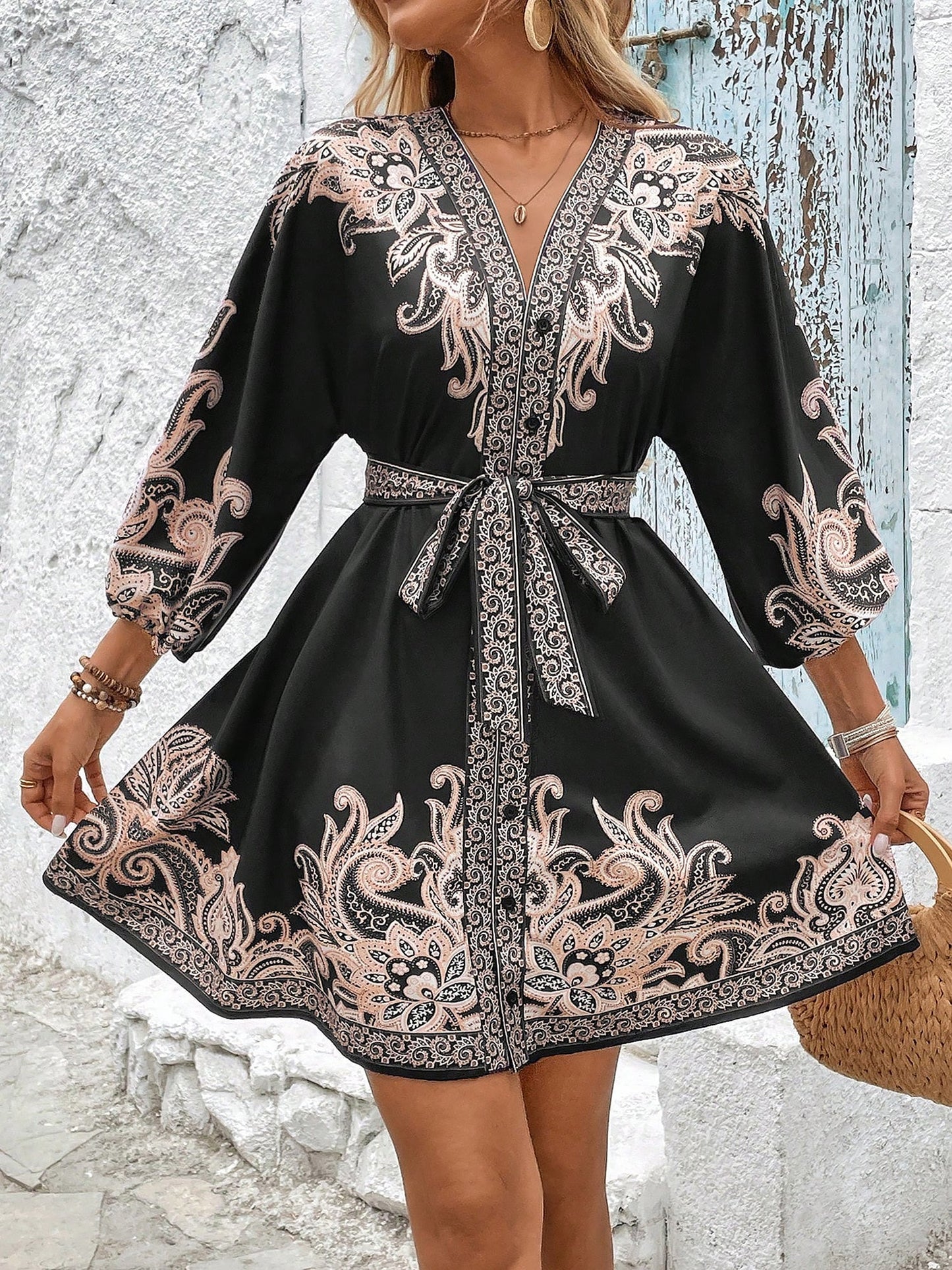 Black - Woman wearing a woven boho V-neck mini shirt dress with ornate paisley print and voluminous sleeves, holding a woven straw bag outdoors.