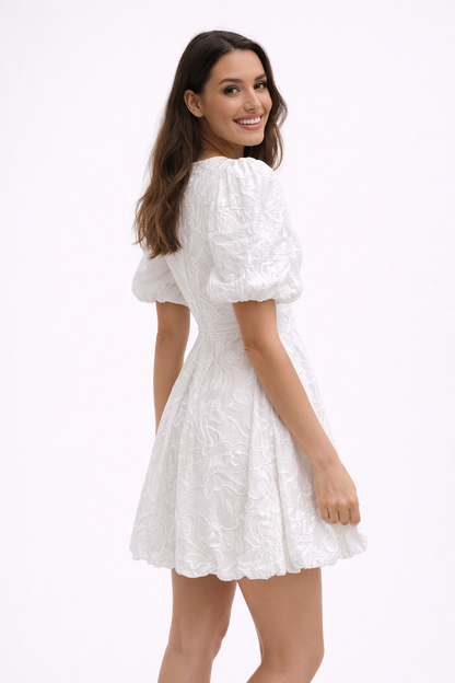 A woman wearing a woven floral A-line mini dress with puff sleeves and a bubble hem, standing against a plain studio background.