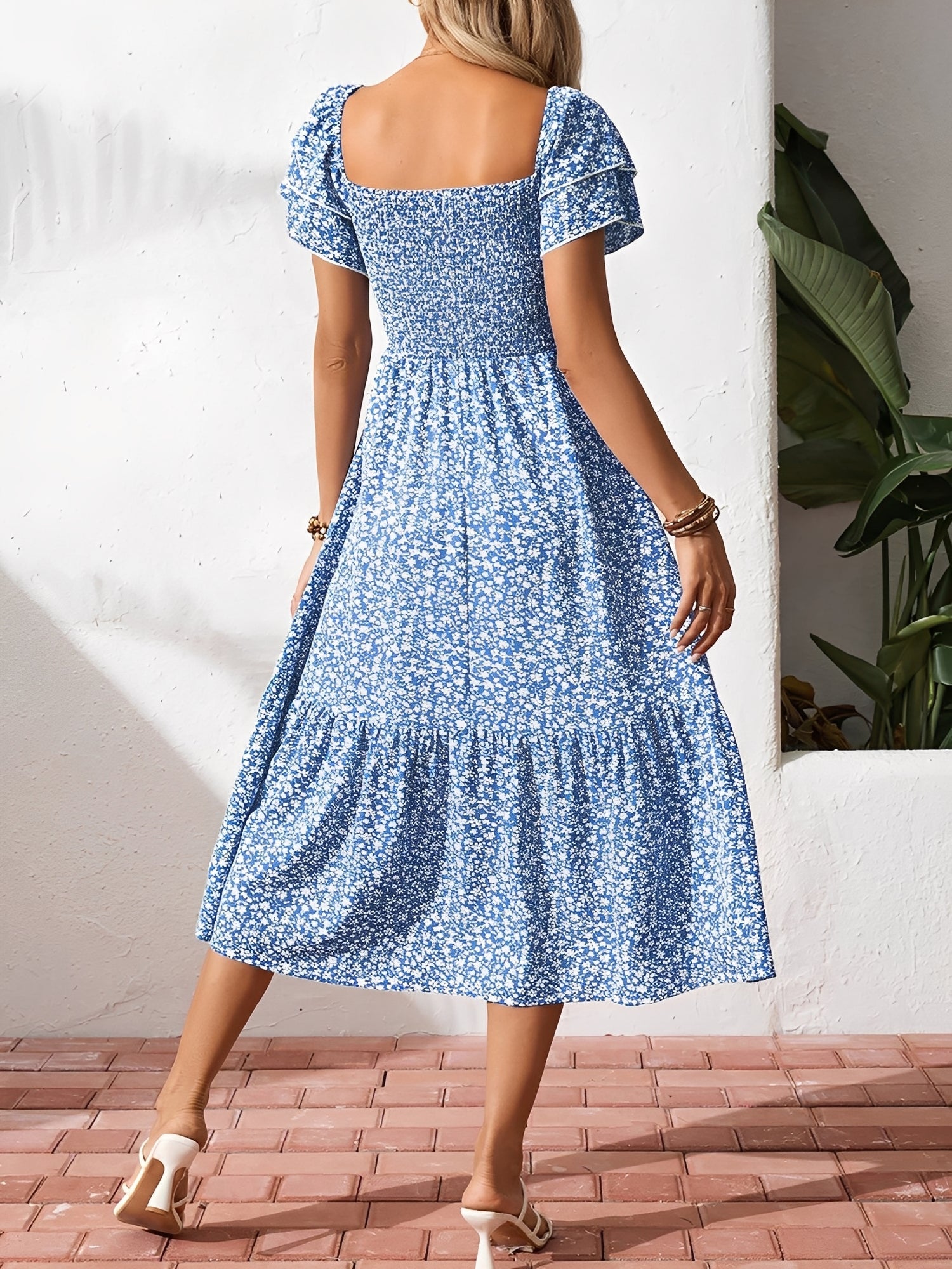 Blue - Woman wearing a woven floral square-neck midi dress with a smocked bodice, flutter sleeves, and a tiered skirt, styled with a straw hat and woven bag.