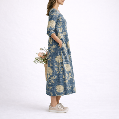 Blue - Woman wearing a woven floral V-neck midi dress with three-quarter sleeves and side pockets, styled with trainers and a straw bag.