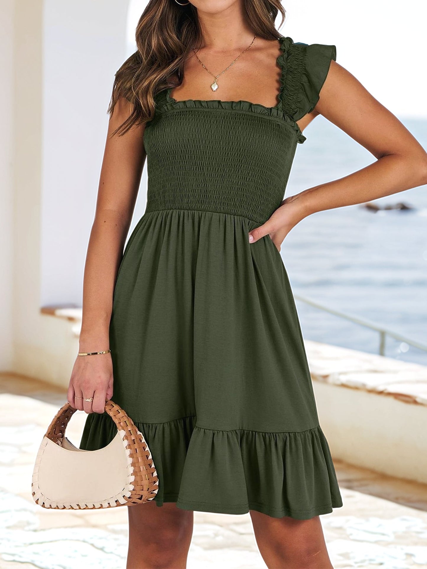 Army Green - Woman wearing a woven smock square-neck mini dress with flutter sleeves and a tiered ruffle hem, standing outdoors by the sea.
