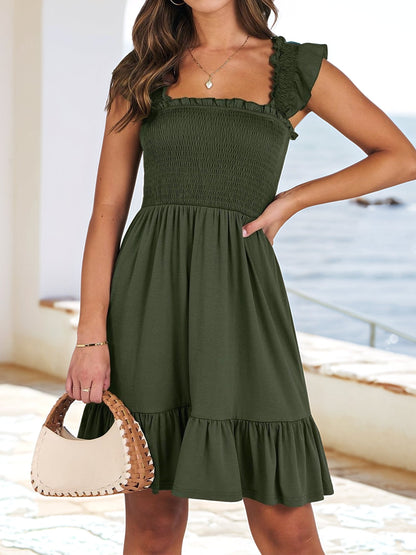 Army Green - Woman wearing a woven smock square-neck mini dress with flutter sleeves and a tiered ruffle hem, standing outdoors by the sea.