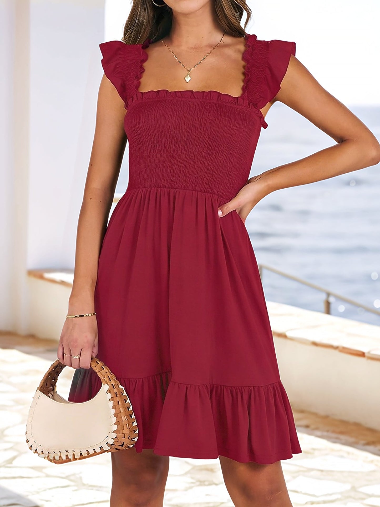 Burgundy - Woman wearing a woven smock square-neck mini dress with flutter sleeves and a tiered ruffle hem, standing outdoors by the sea.