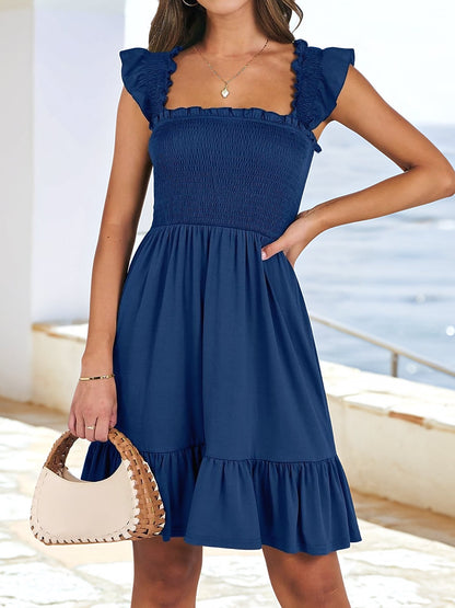 Navy Blue - Woman wearing a woven smock square-neck mini dress with flutter sleeves and a tiered ruffle hem, standing outdoors by the sea.