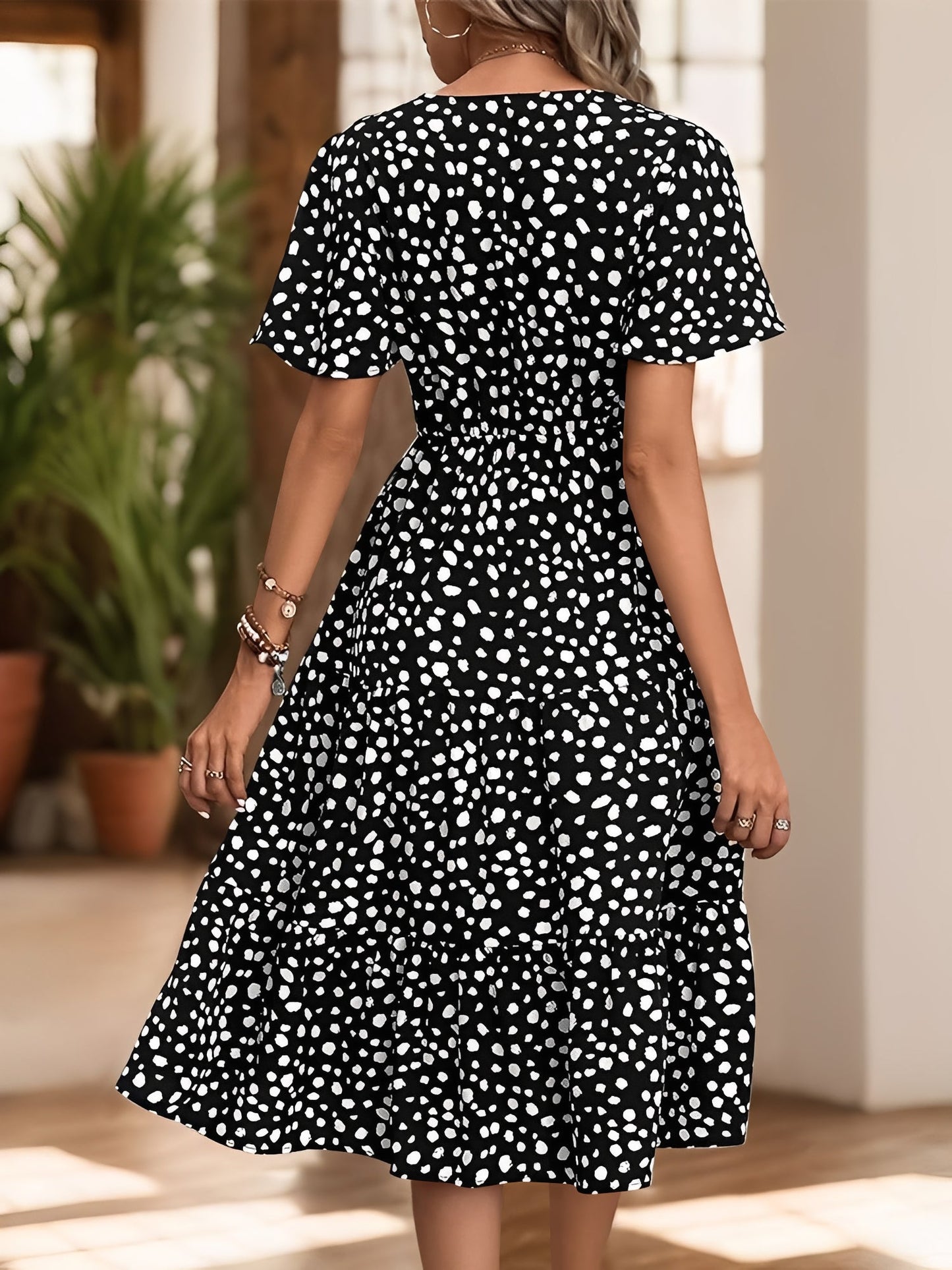 Black - Woman wearing a spot pattern V-neck tiered midi dress with short sleeves and side pockets, standing in an architectural hallway.