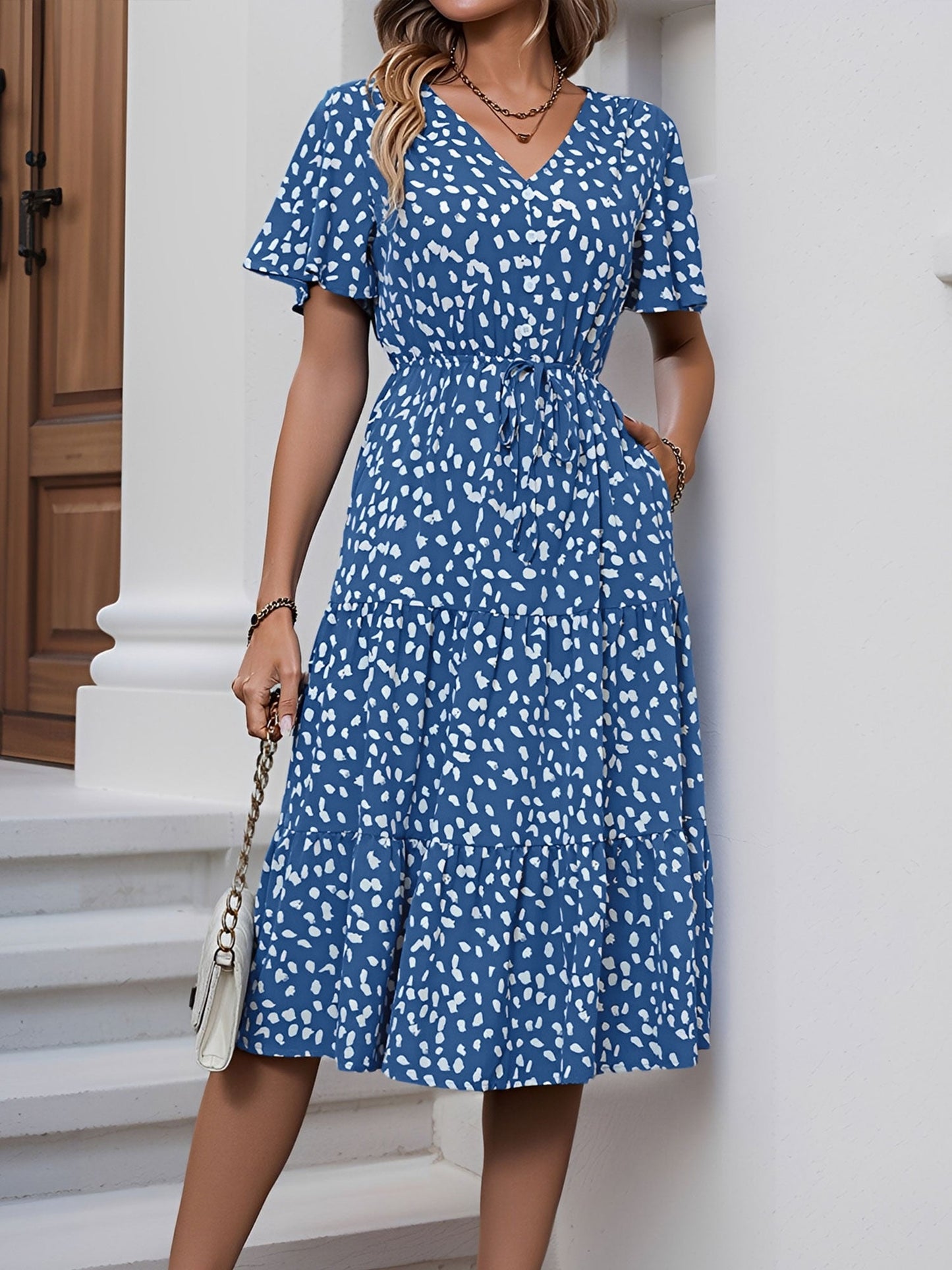 Blue - Woman wearing a spot pattern V-neck tiered midi dress with short sleeves and side pockets, standing in an architectural hallway.