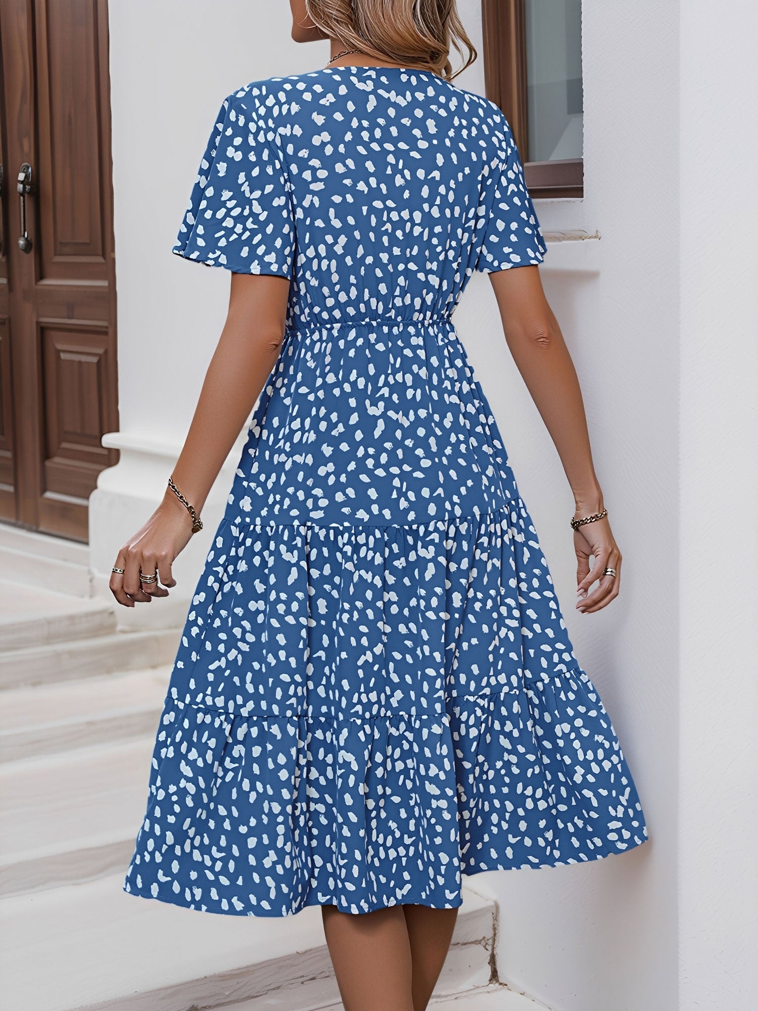 Blue - Woman wearing a spot pattern V-neck tiered midi dress with short sleeves and side pockets, standing in an architectural hallway.