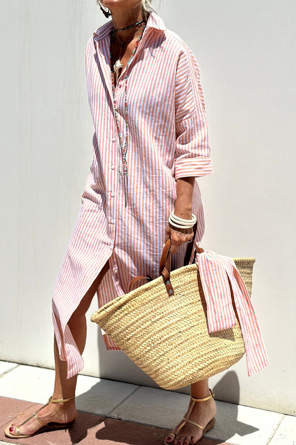 Pink - A woman wearing a striped, button-down midi-length shirt dress with 3/4 sleeves, carrying a woven straw basket bag outdoors.