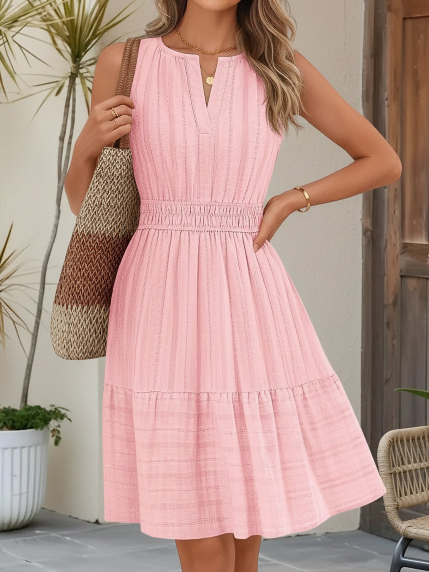 Pink - A woman wearing a sleeveless, textured woven midi dress with a notched V-neckline, shirred waist, and tiered skirt, holding a woven tote bag in a bright outdoor setting.