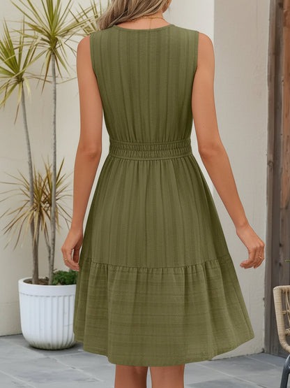 Olive Green - A woman wearing a sleeveless, textured woven midi dress with a notched V-neckline, shirred waist, and tiered skirt, holding a woven tote bag in a bright outdoor setting.