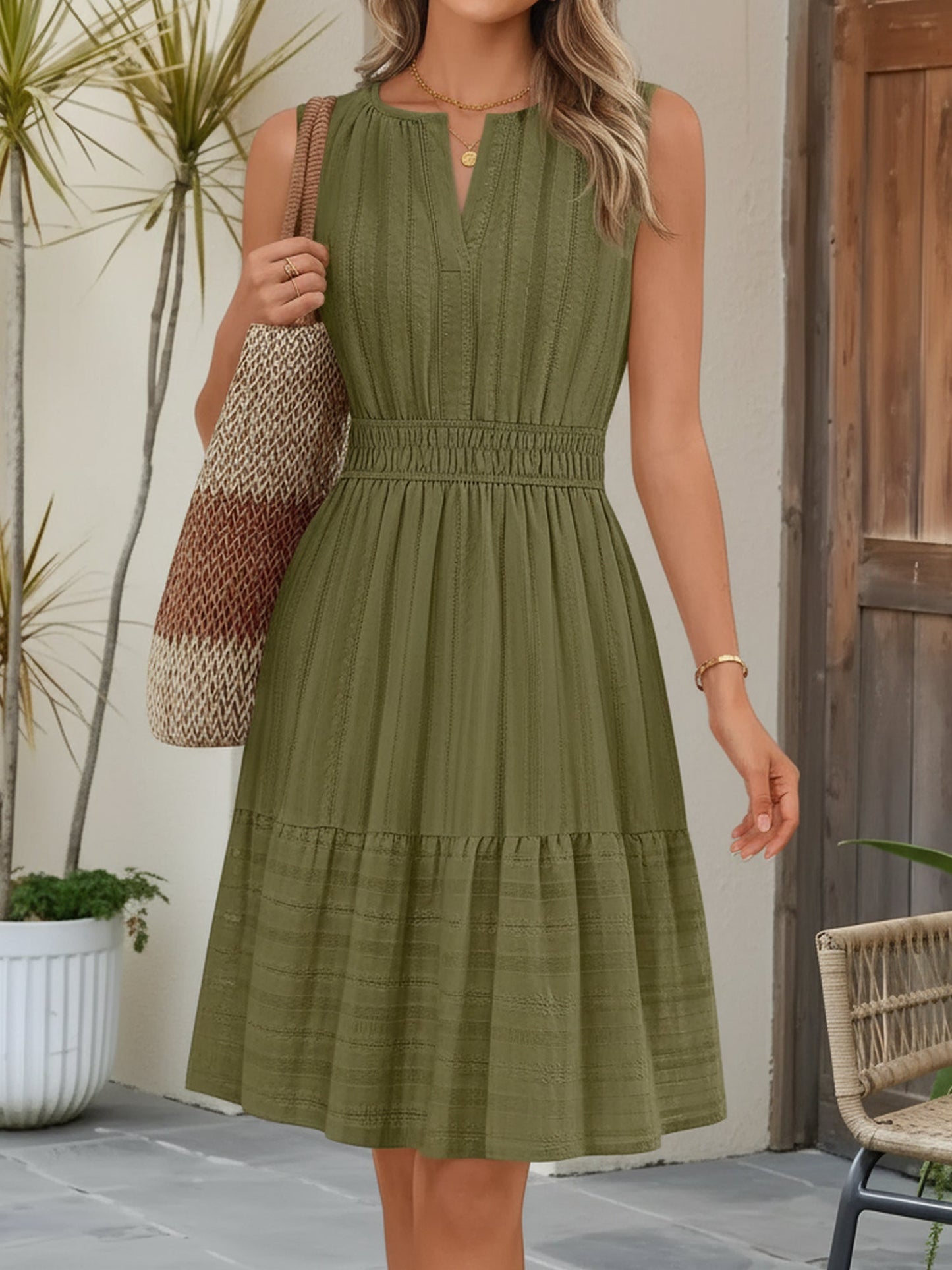Olive Green - A woman wearing a sleeveless, textured woven midi dress with a notched V-neckline, shirred waist, and tiered skirt, holding a woven tote bag in a bright outdoor setting.