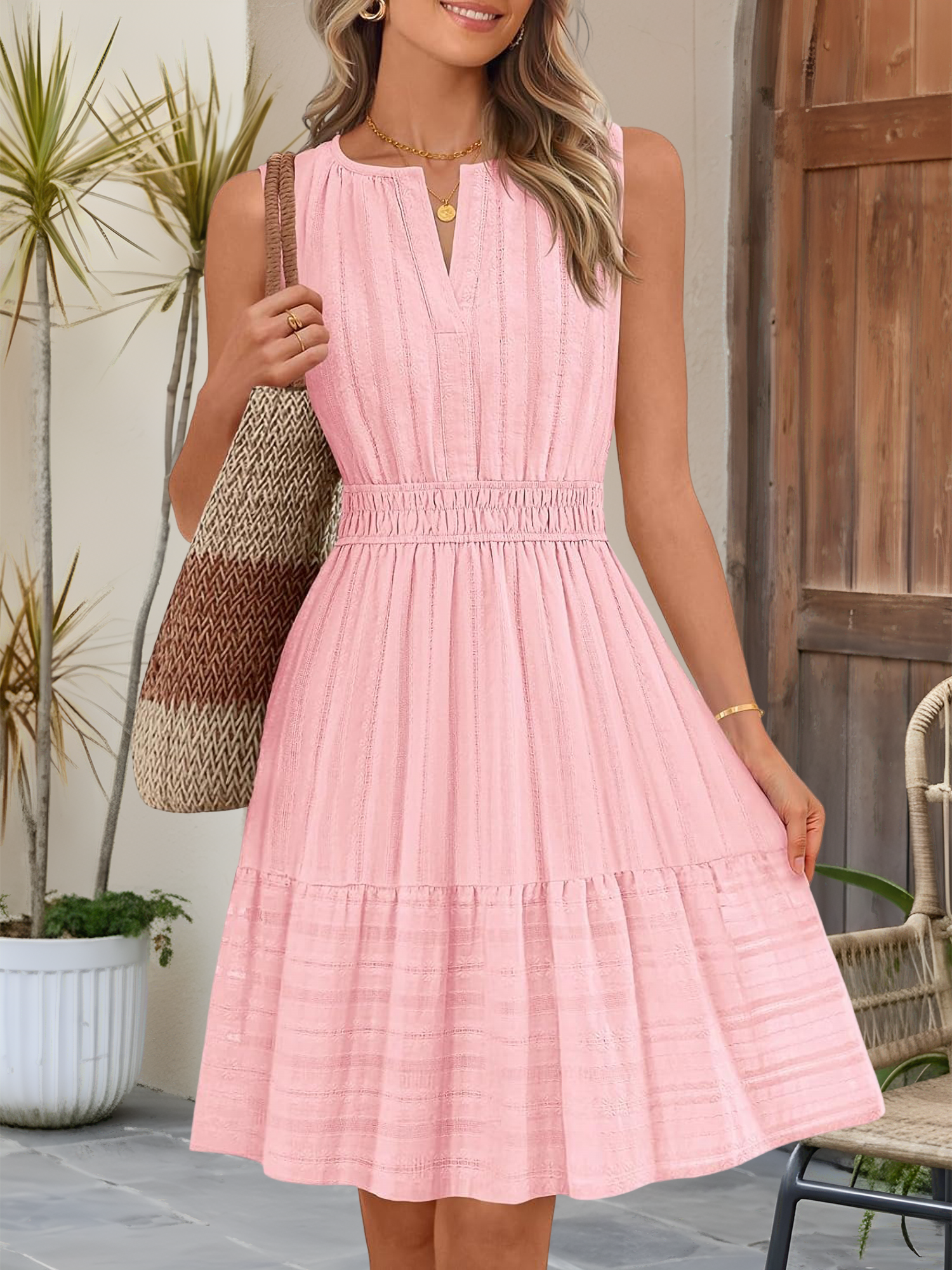 Pink - A woman wearing a sleeveless, textured woven midi dress with a notched V-neckline, shirred waist, and tiered skirt, holding a woven tote bag in a bright outdoor setting.