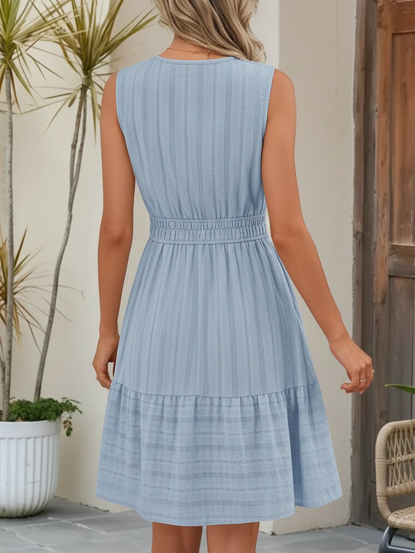 Blue - A woman wearing a sleeveless, textured woven midi dress with a notched V-neckline, shirred waist, and tiered skirt, holding a woven tote bag in a bright outdoor setting.