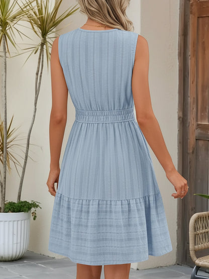 Blue - A woman wearing a sleeveless, textured woven midi dress with a notched V-neckline, shirred waist, and tiered skirt, holding a woven tote bag in a bright outdoor setting.