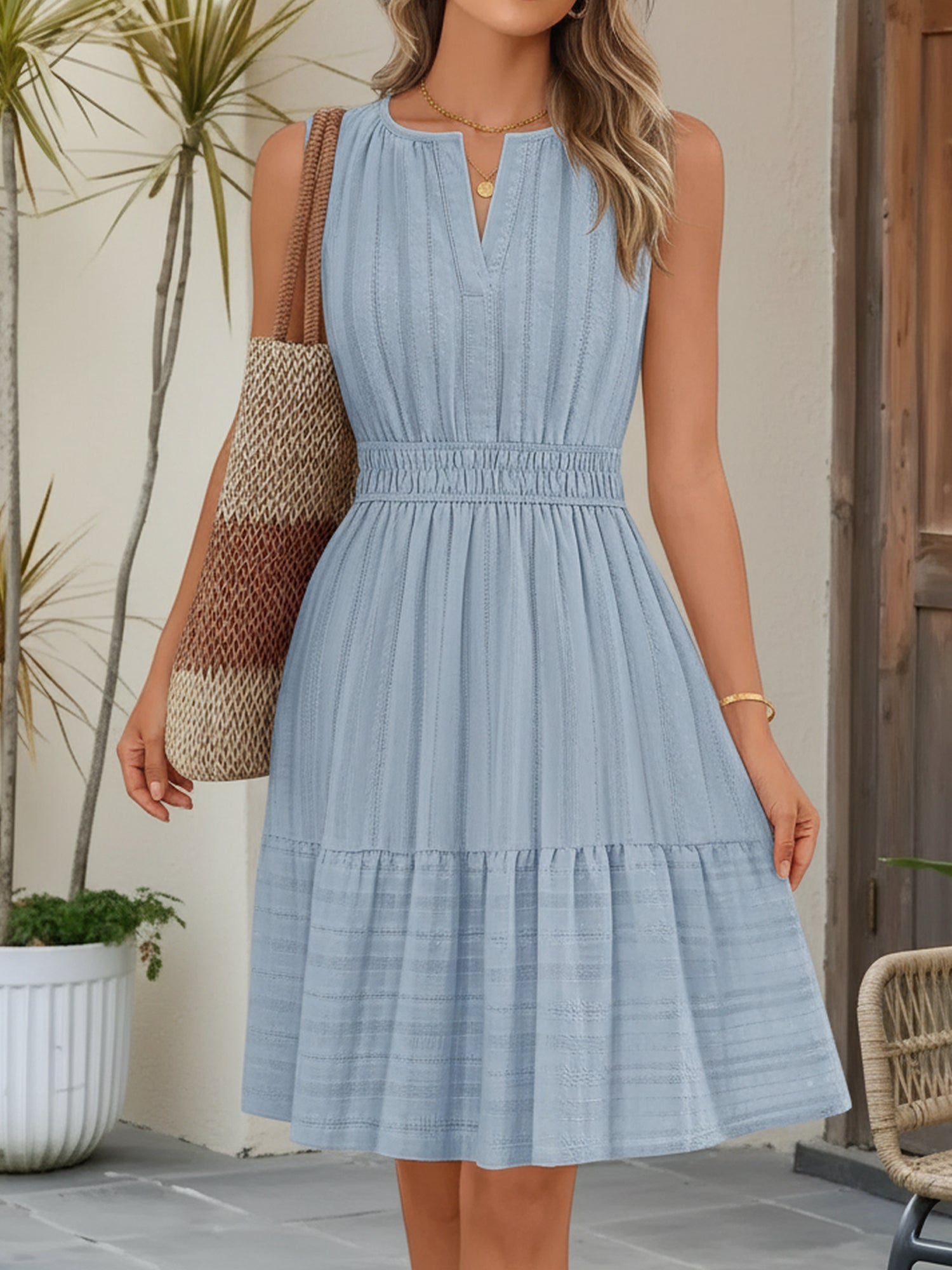 Blue - A woman wearing a sleeveless, textured woven midi dress with a notched V-neckline, shirred waist, and tiered skirt, holding a woven tote bag in a bright outdoor setting.