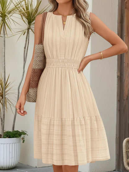 Ivory - A woman wearing a sleeveless, textured woven midi dress with a notched V-neckline, shirred waist, and tiered skirt, holding a woven tote bag in a bright outdoor setting.